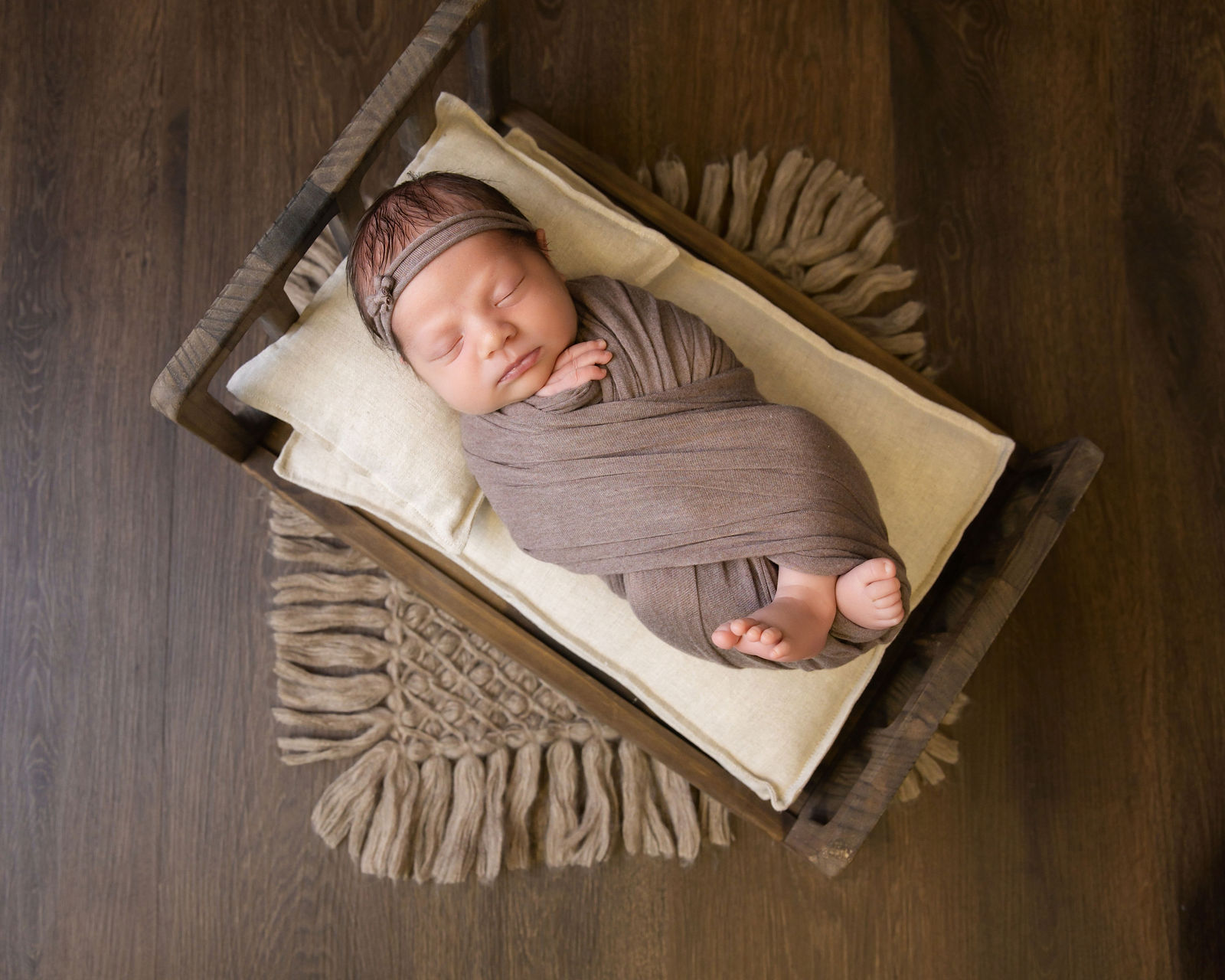 Sleeping newborn wrapped in brown fabric and posed in a small wooden bed with neutral styling