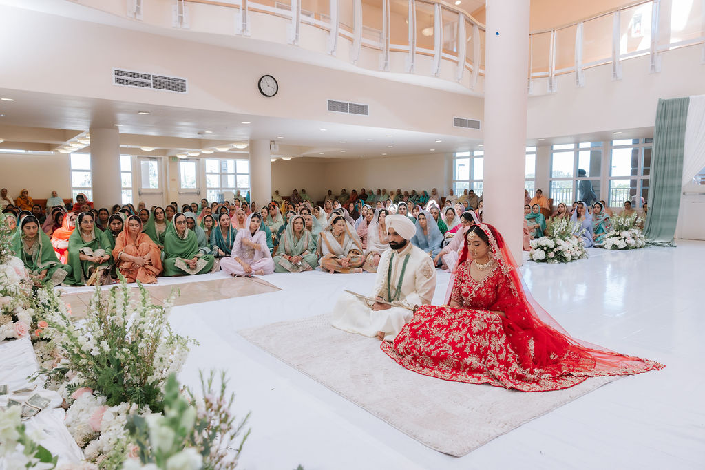 Mandeep and Naveet seated for Anand Karaj with congregation, tall columns, windows and the balcony visible across the hall