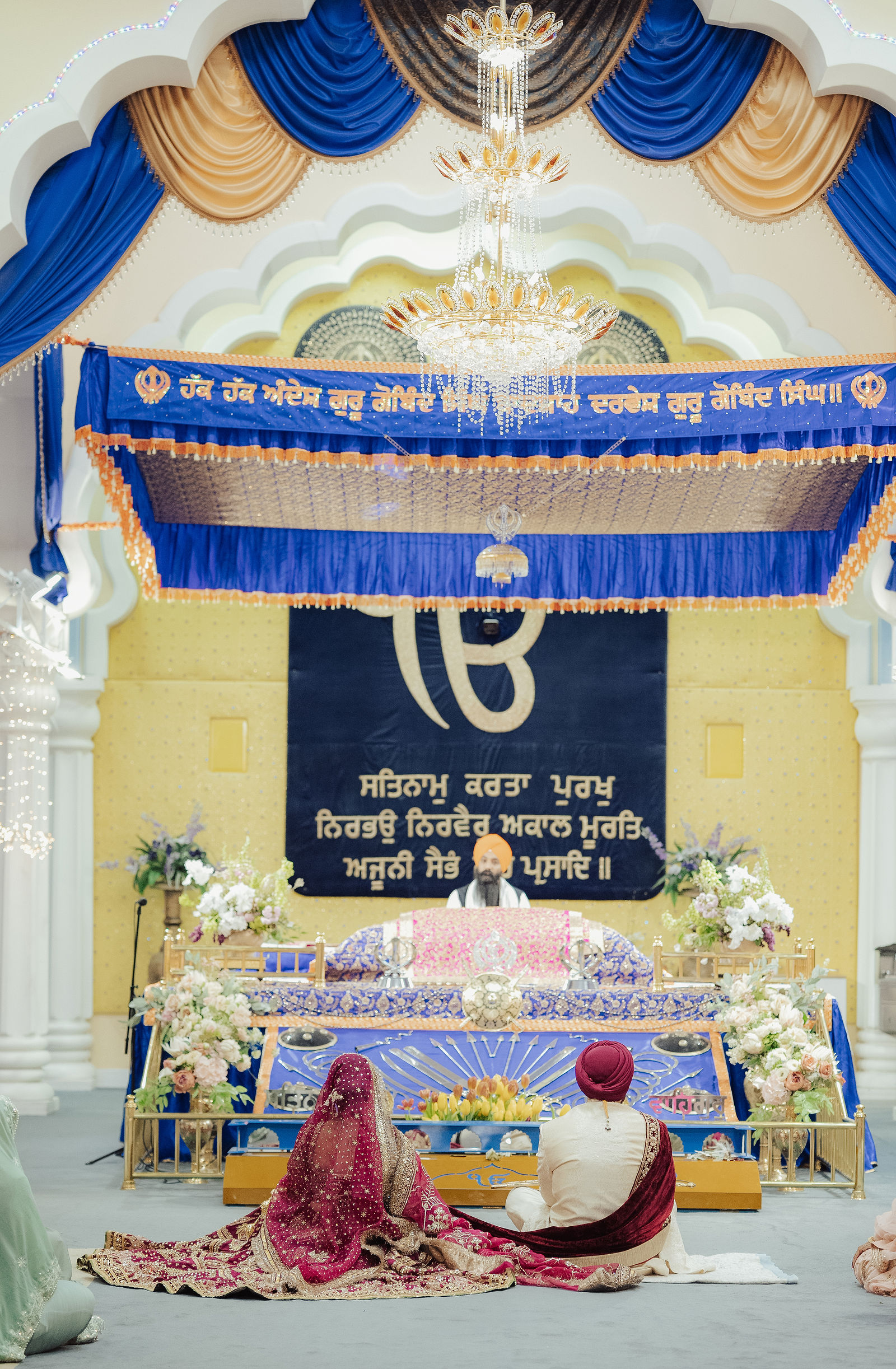 Gurleen and Jaiprit seated during their Anand Karaj at San Jose Gurdwara beneath blue-and-gold draping, chandeliers and white arches.