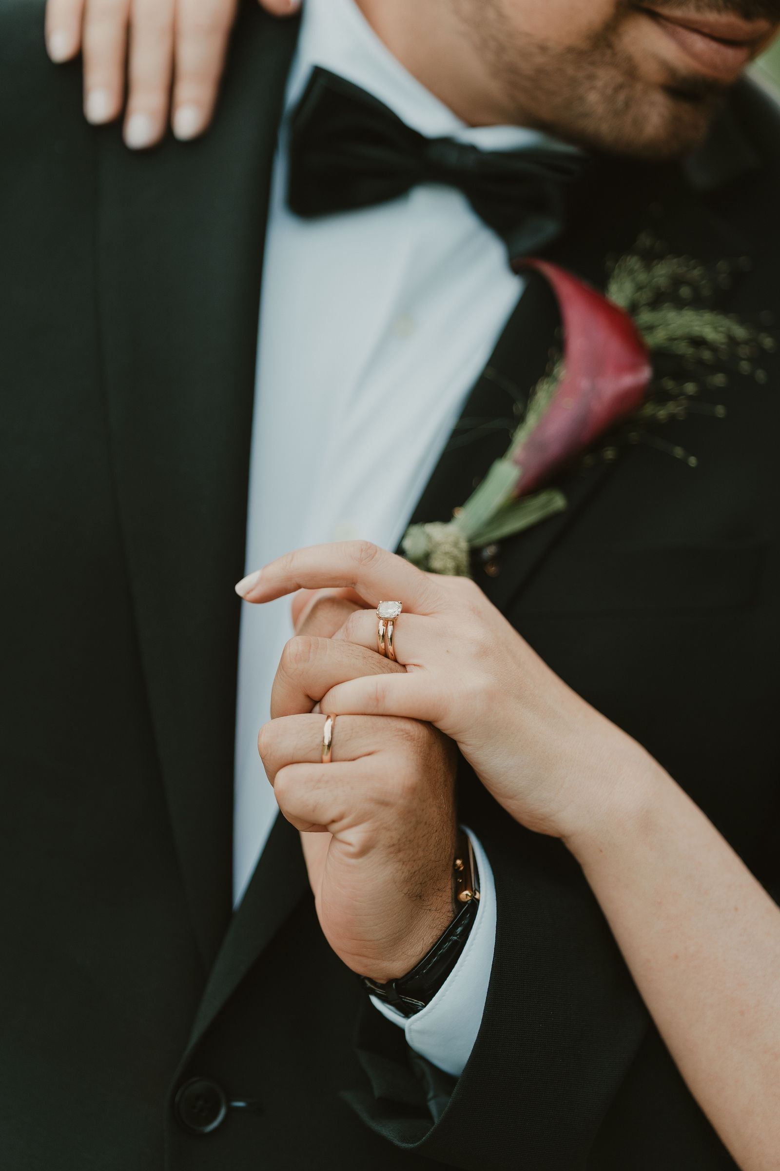 The couple’s hands and rings against a dark suit and burgundy flower
