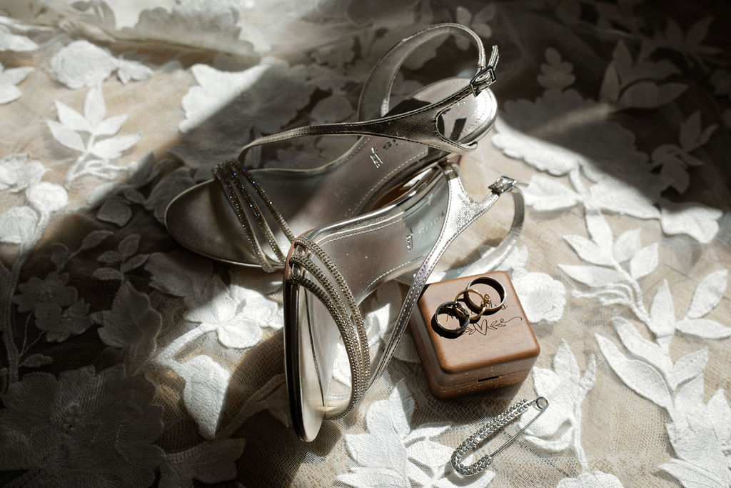 Chaussures argentées et alliances posées sur la dentelle de la robe de mariée.
