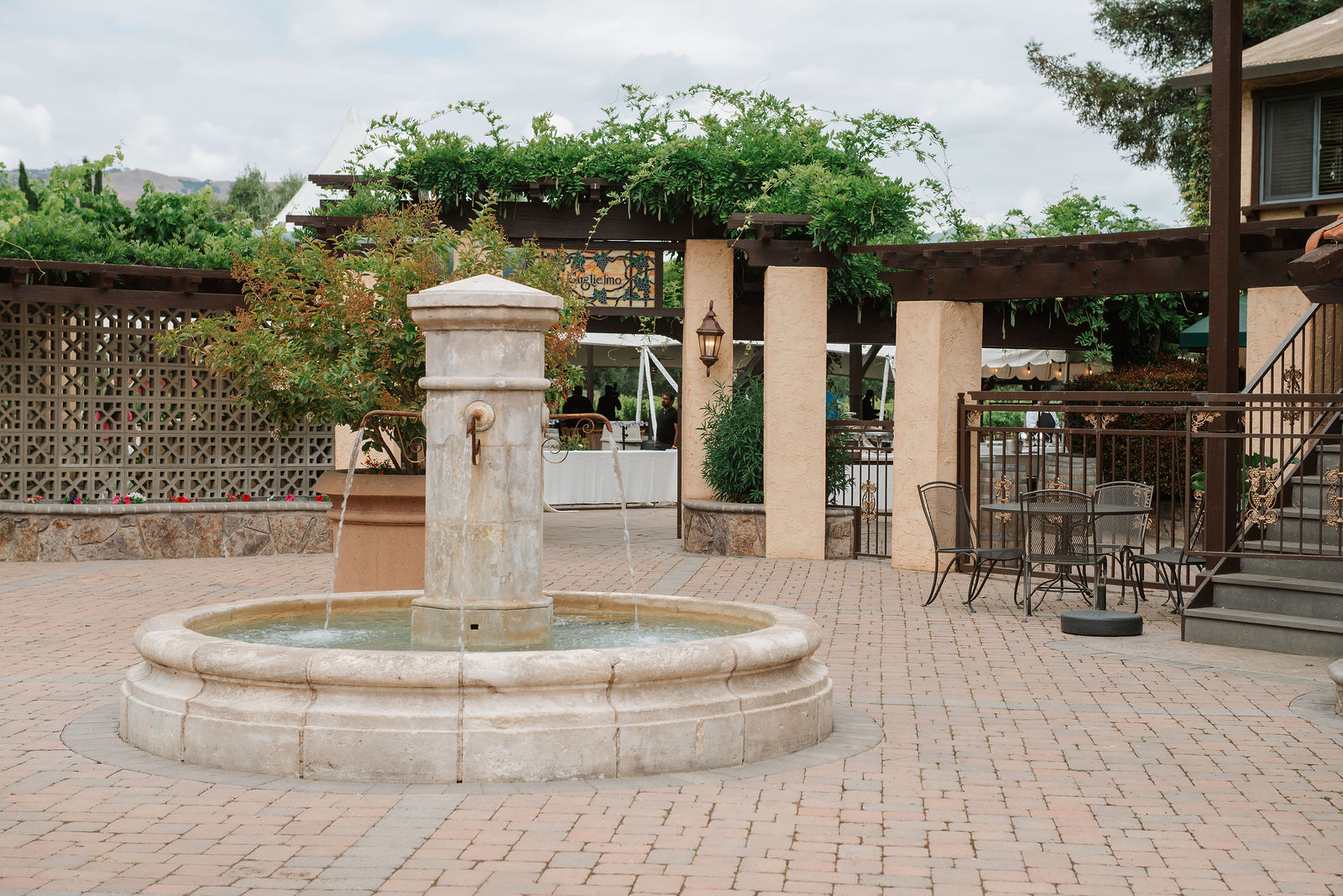 A fountain stands in Guglielmo Winery’s brick courtyard, with a vine-covered pavilion and the winery sign behind it.