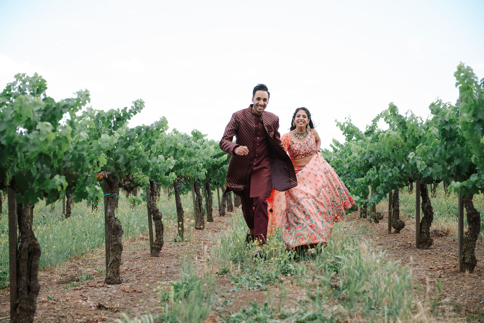 Rishi and Nanki smile while moving together between rows of grapevines.