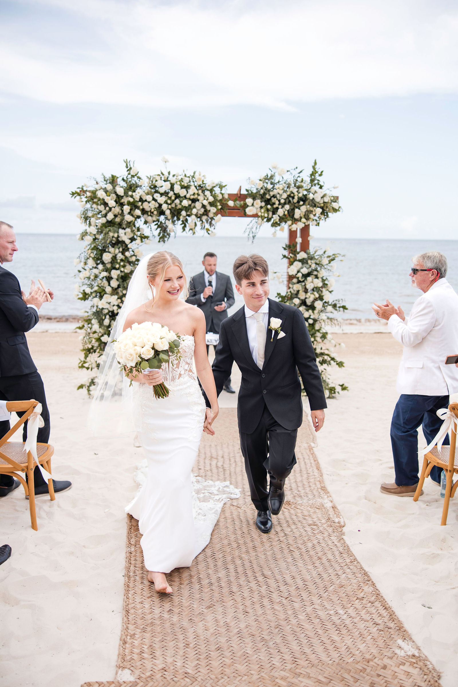 A couple walking back down the aisle of a beach ceremony, the Caribbean behind them and a floral arch overhead