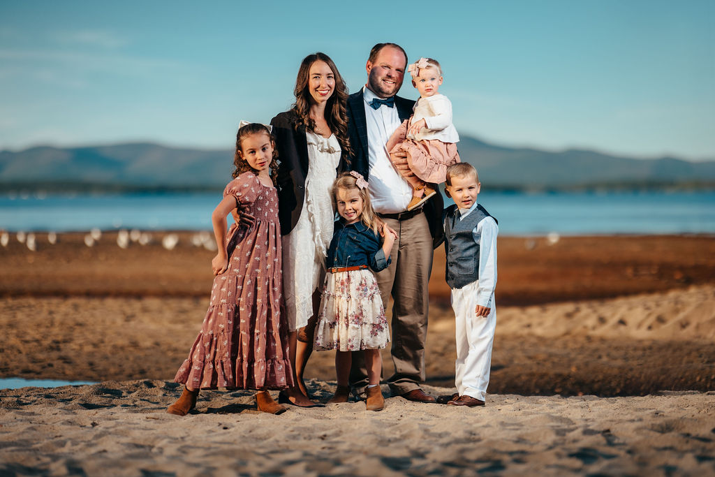Haynes family of six posed on a sandy beach at golden hour with a lake and mountains behind them