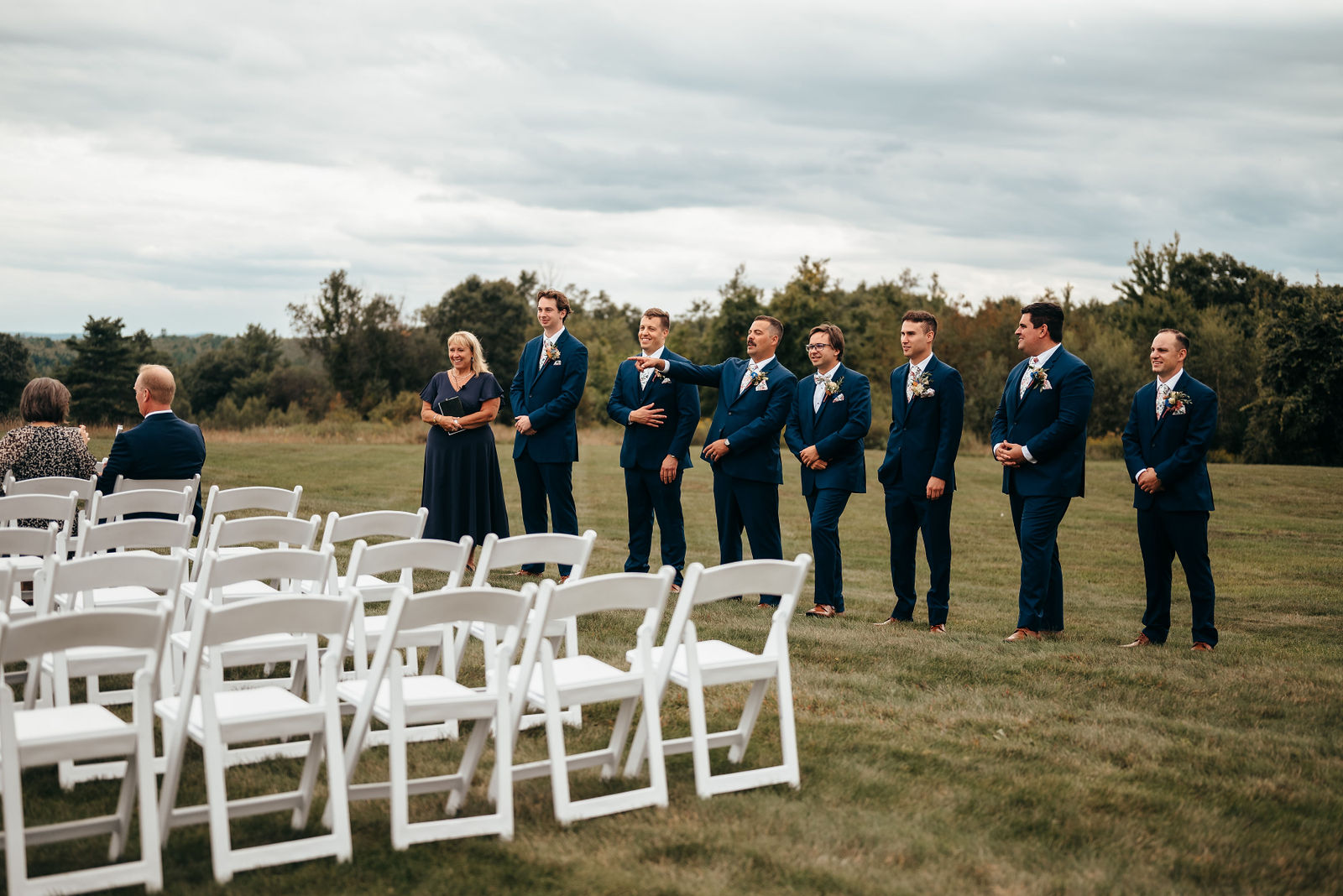 Groomsmen in navy suits lined up behind white ceremony chairs on the lawn under a dramatic sky at Dell-Lea