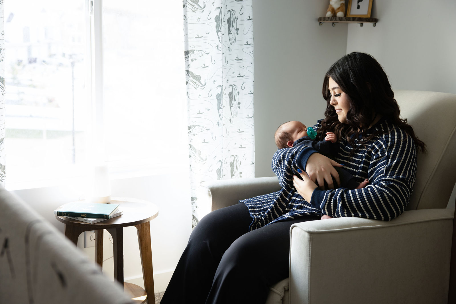 Mother sitting beside a bright nursery window while holding her newborn baby in her arms