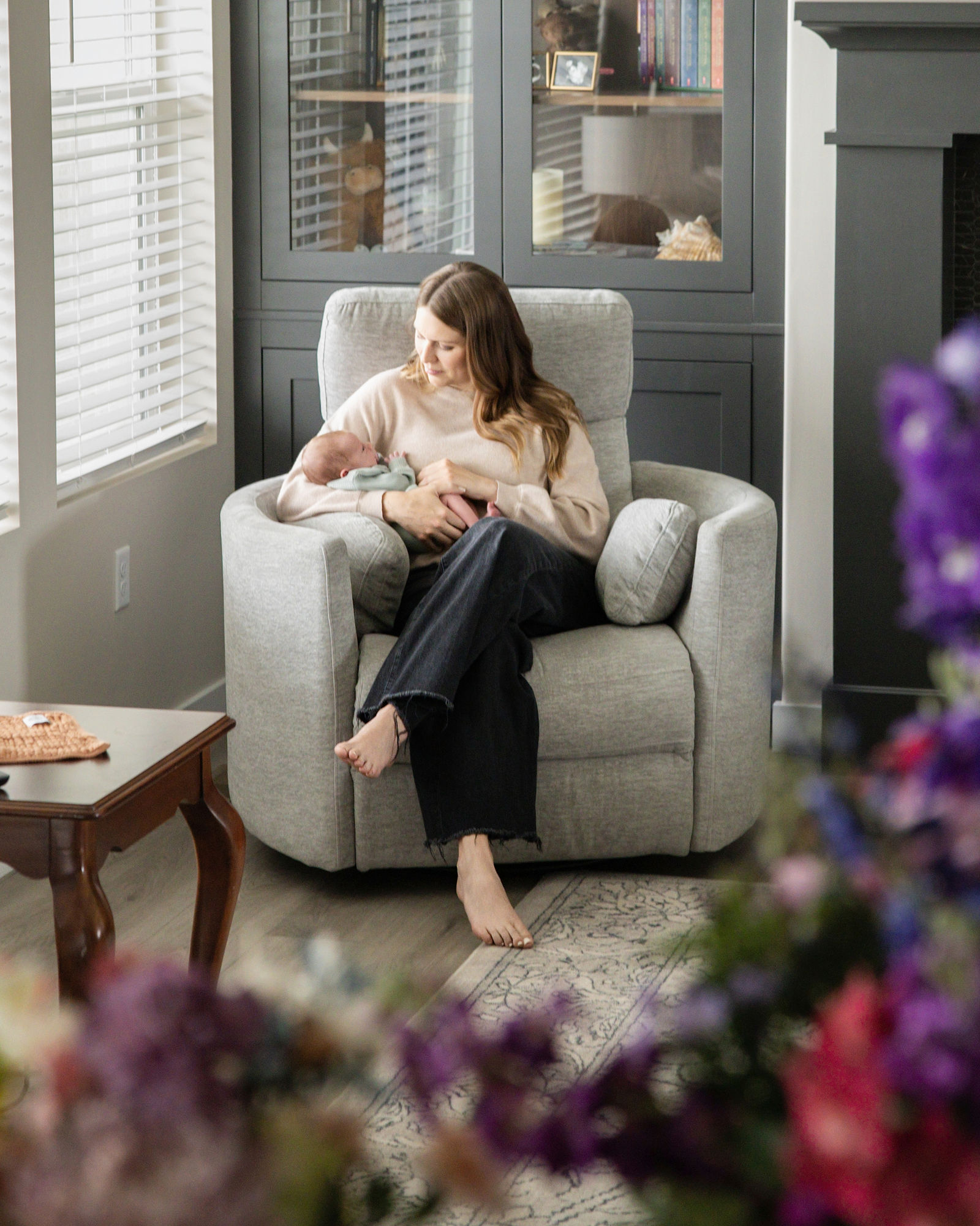 Mother holding her newborn baby in a chair beside a bright window during a lifestyle newborn session