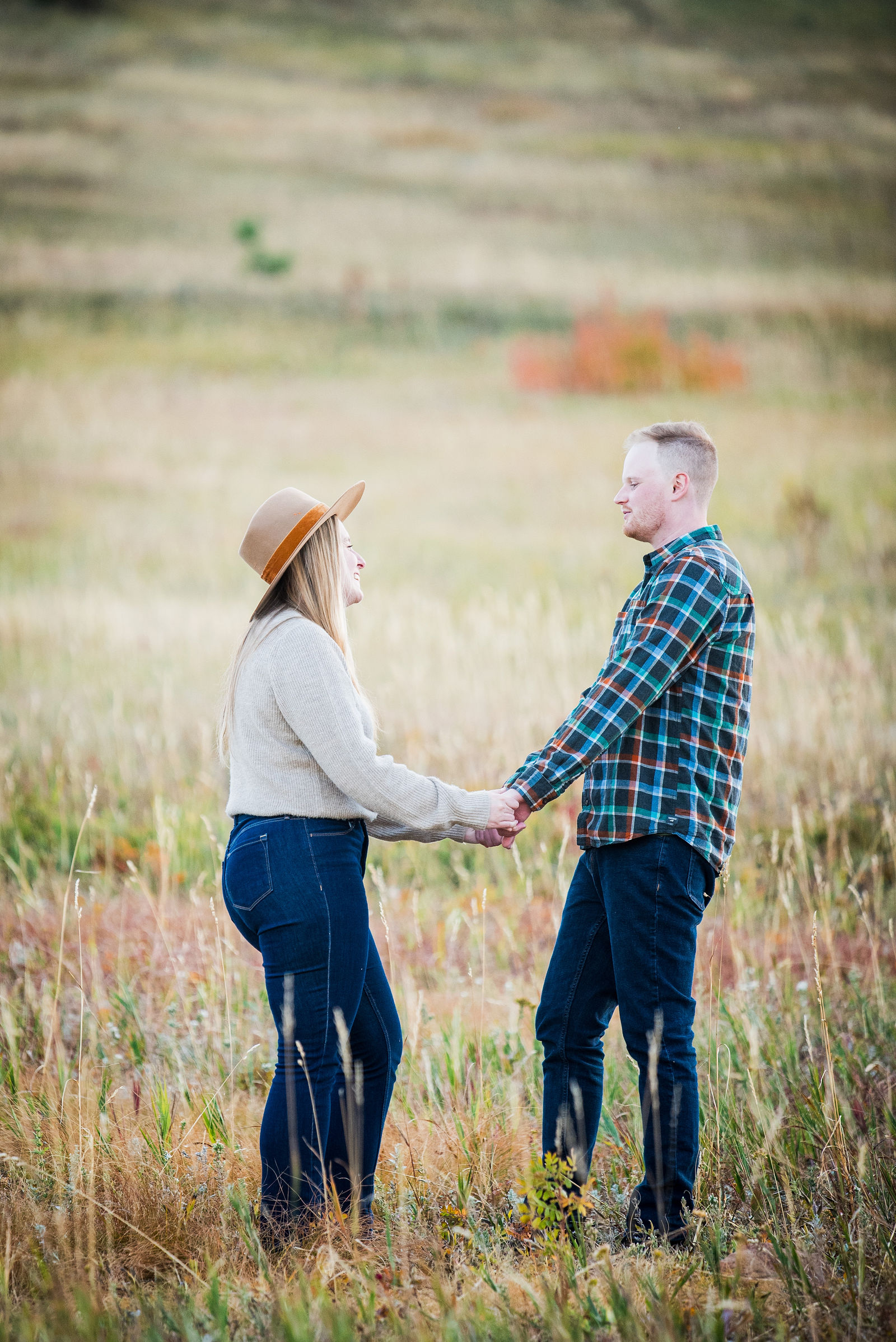 Engaged couple in a cream sweater, dark jeans and a tan felt hat beside a navy plaid flannel in the Boulder foothills