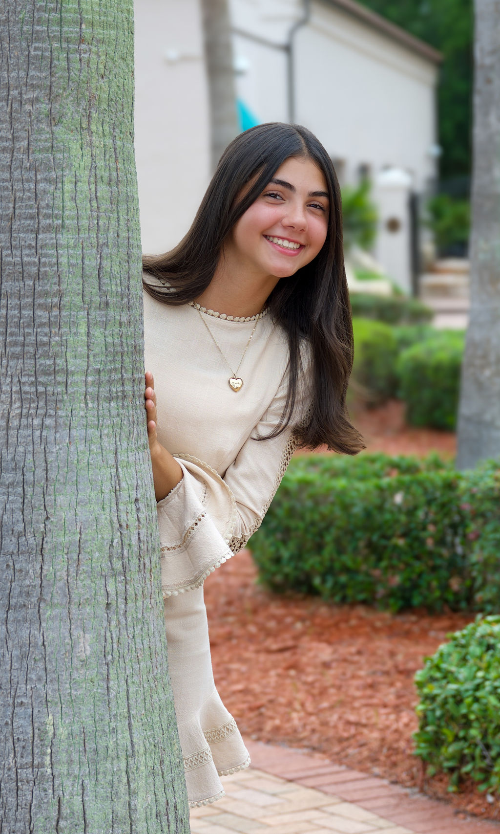 Young girl in a cream dress peeking playfully from behind a tree