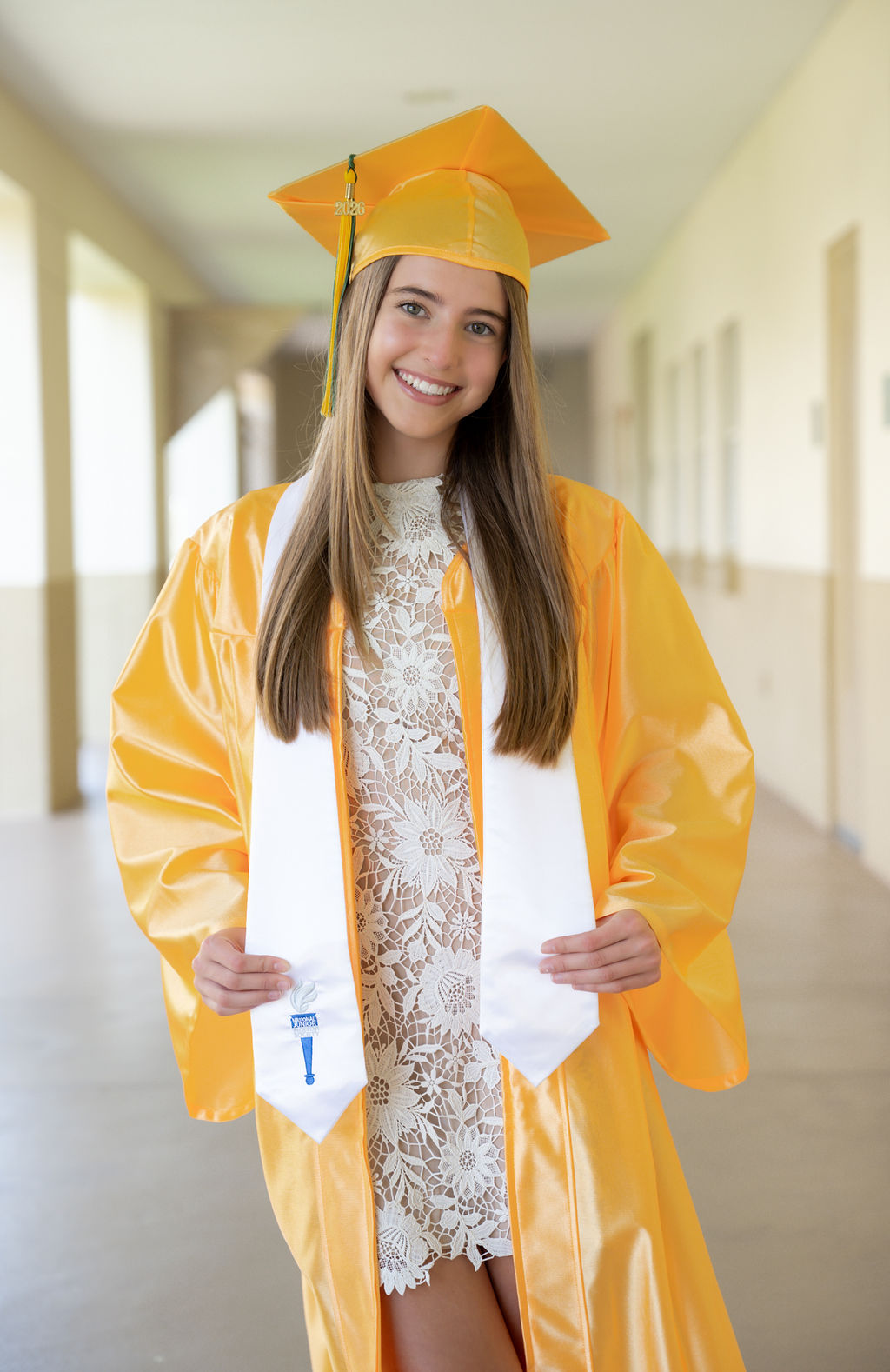 Graduate in a gold cap and gown standing in a bright architectural corridor