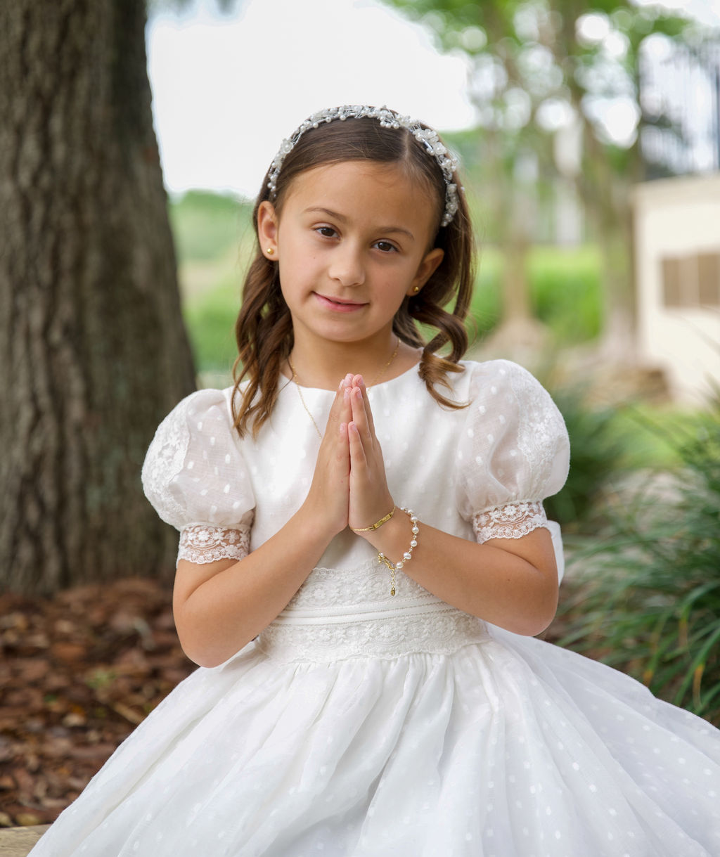 Child in a white communion dress with hands in prayer beside a tree