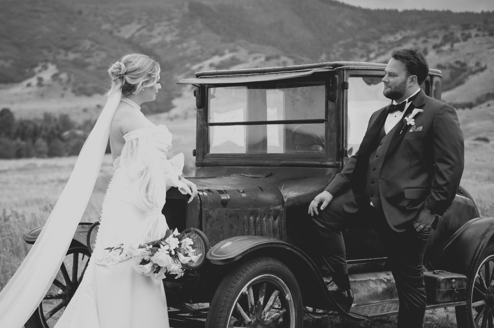Groom leaning against a vintage car in a black tuxedo