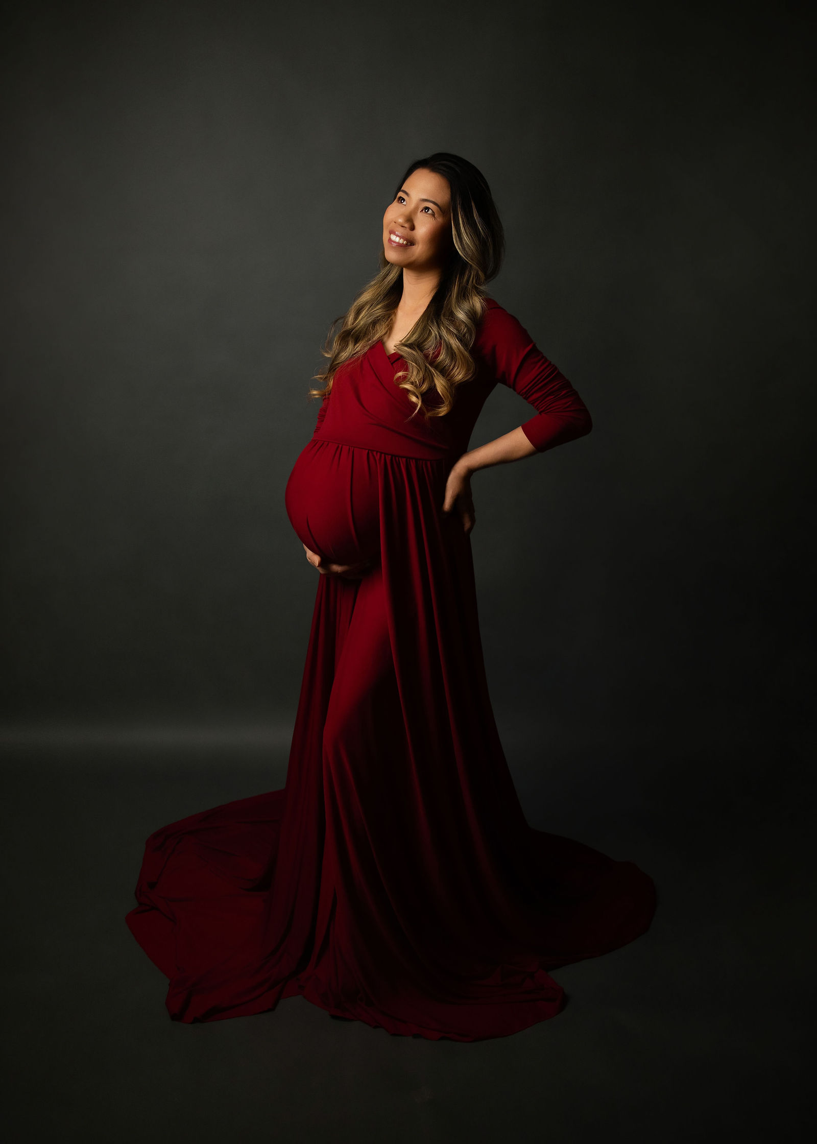 Pregnant woman in a burgundy maternity gown photographed with dramatic studio lighting