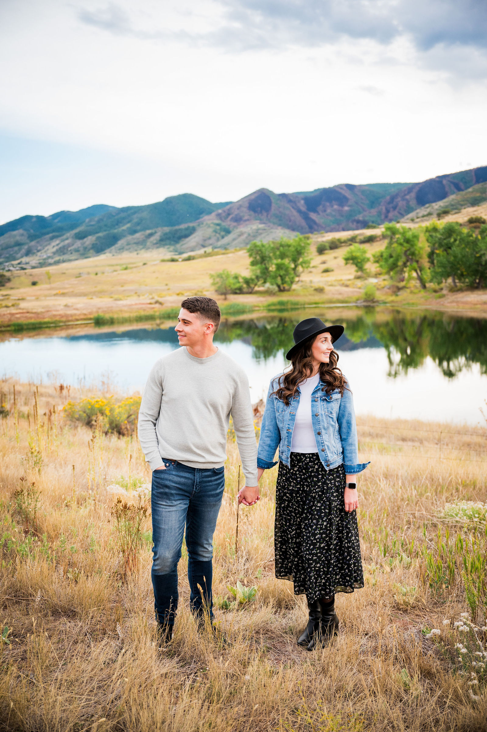 Engaged couple with the woman in a black floral midi skirt, white tee, black felt hat and denim jacket in a Colorado field