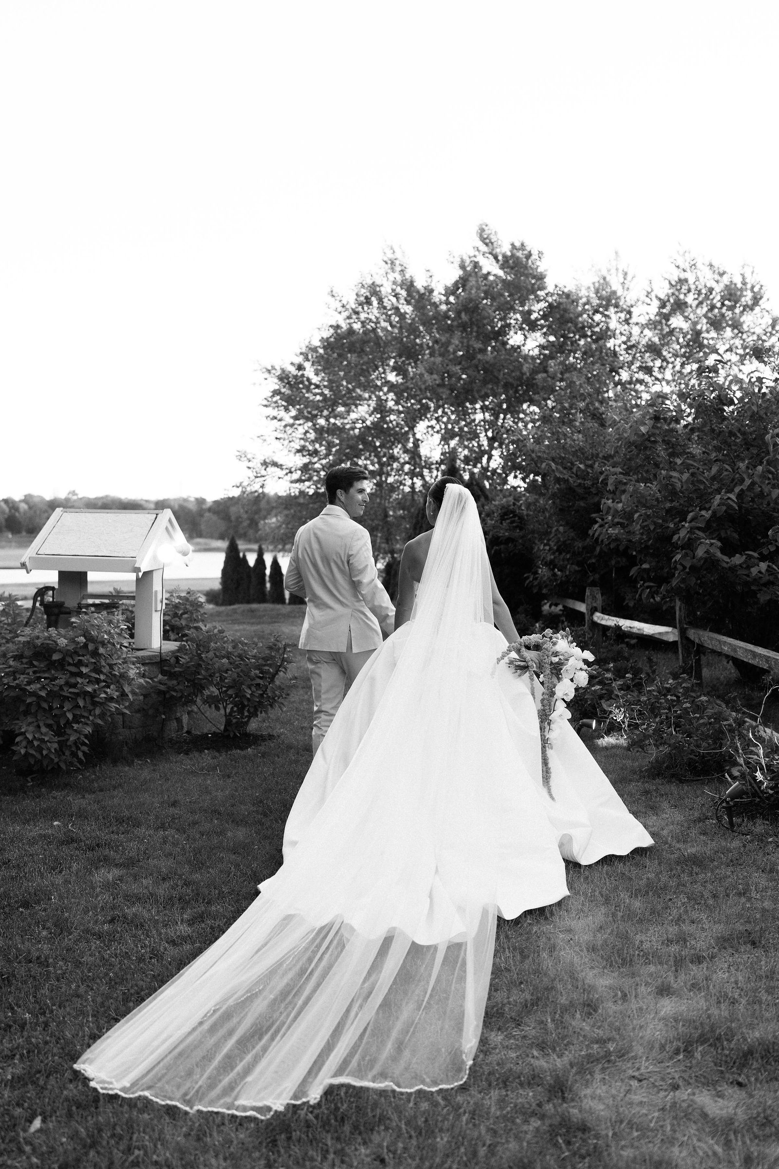 Black-and-white portrait of Andy and Lauren walking across the lawn with Lauren’s veil spread behind her