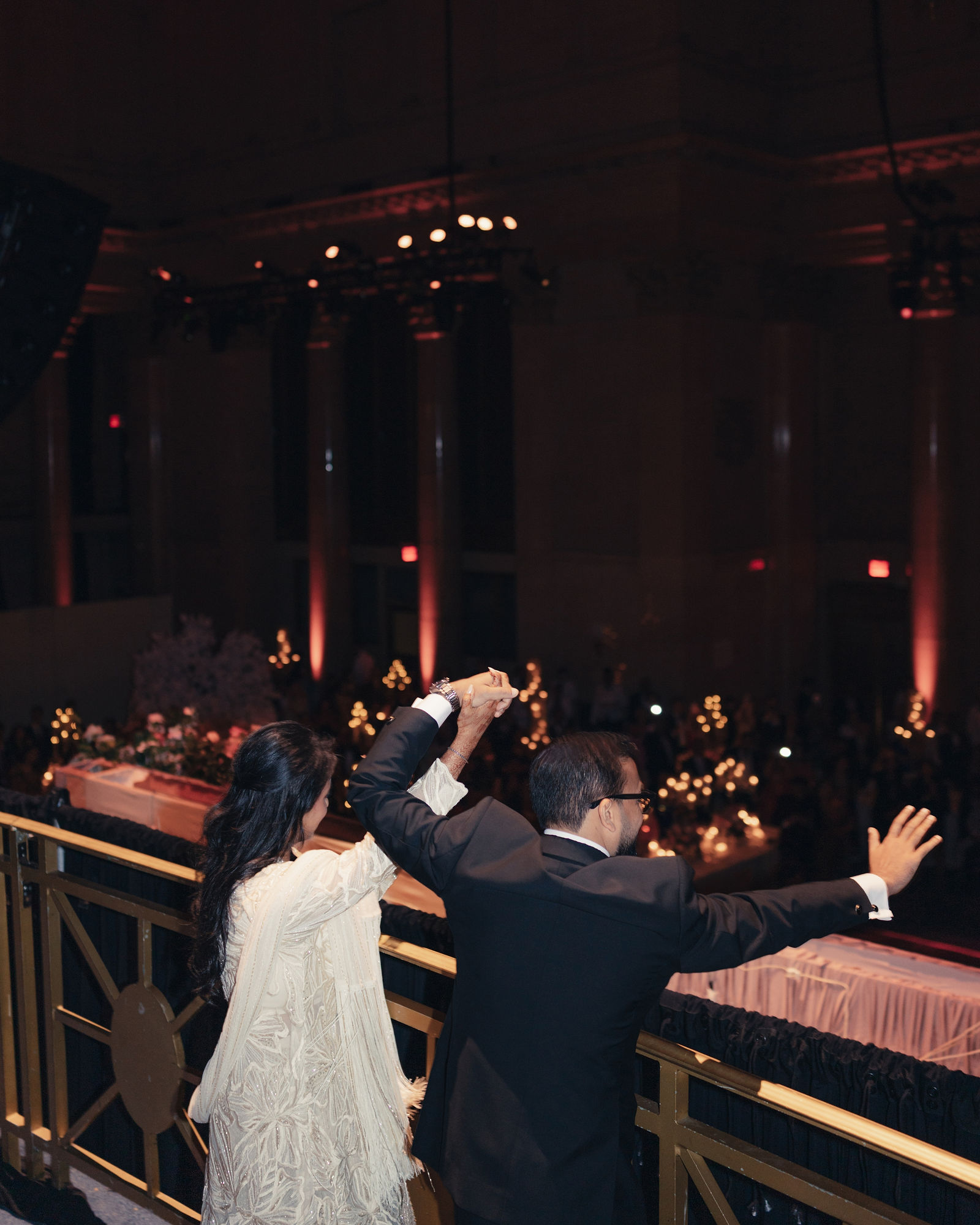 Seen from behind, Anjali and Aditya raise their joined hands and wave from a balcony toward the reception below.