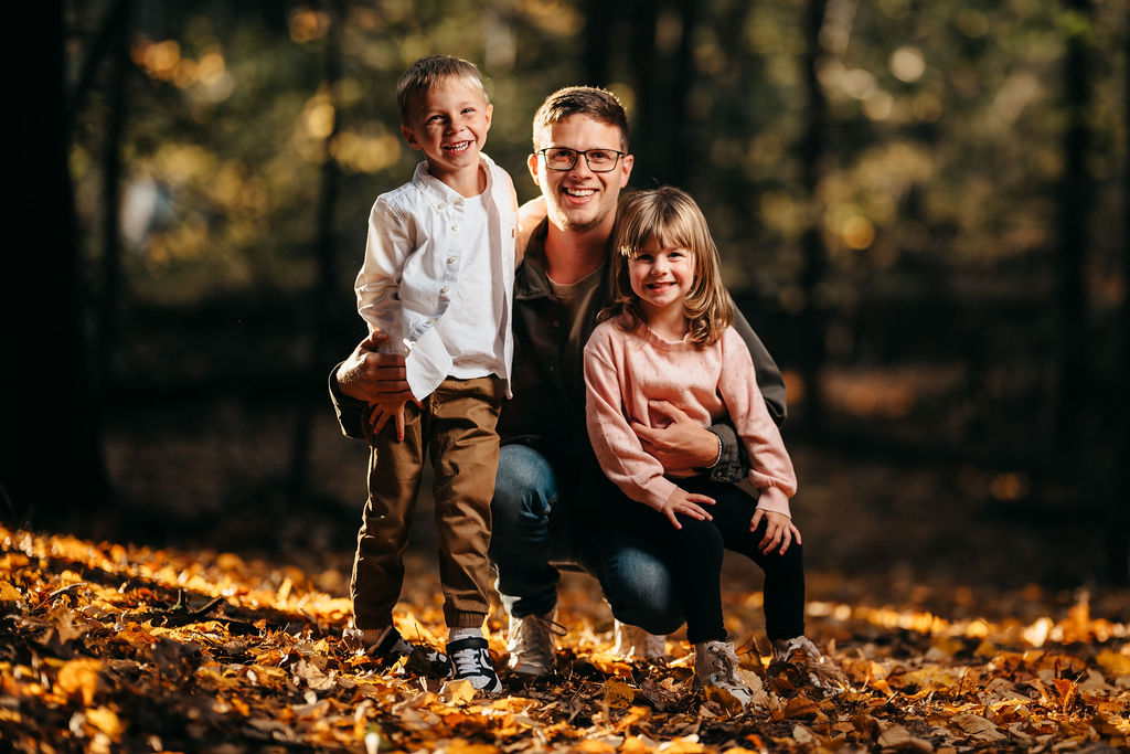 Dad kneeling with his son and daughter on a carpet of golden-orange fallen leaves in backlit woods