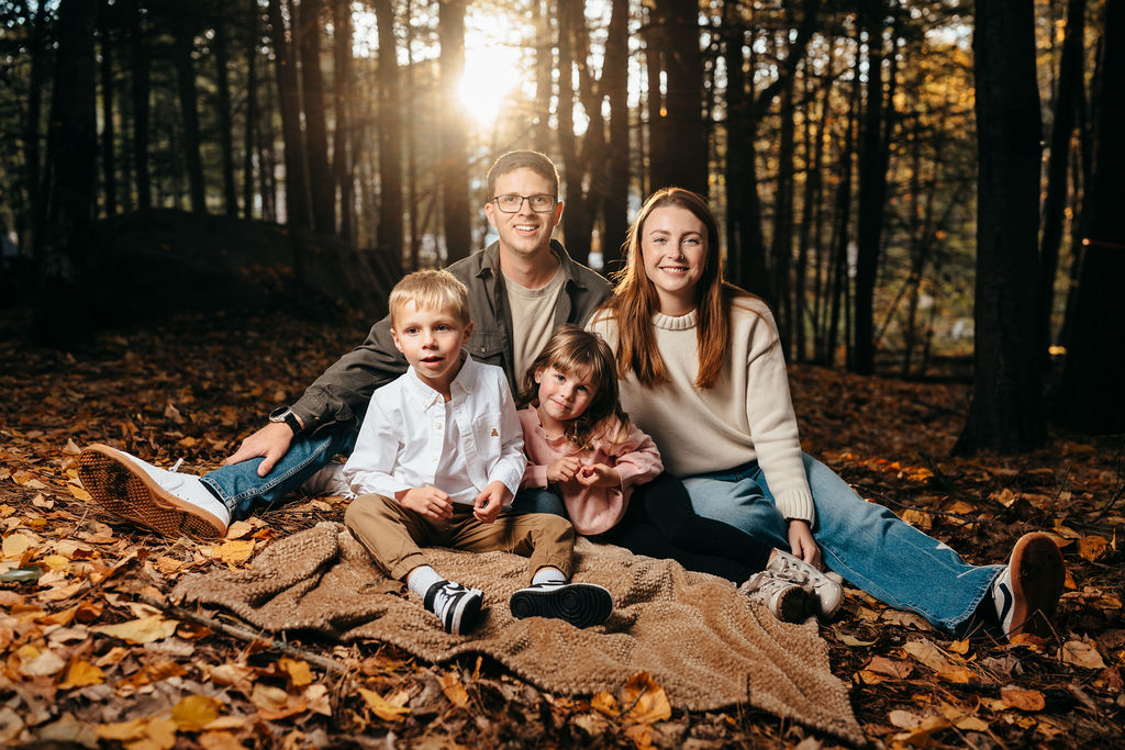 Family of four sitting on a blanket in the woods with sun flare streaming through the trees
