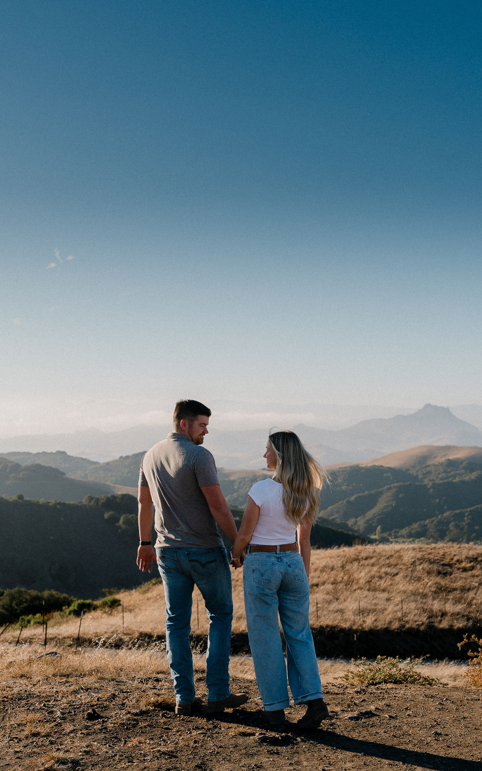 Engaged couple holding hands looking out over golden hills at sunset, Central Coast pre-wedding session
