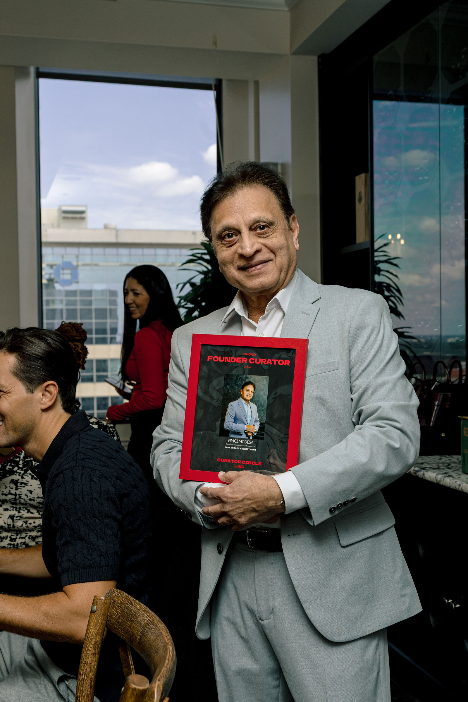 Vincent Desai with his framed Founder Curator plaque, the city in the window behind him.