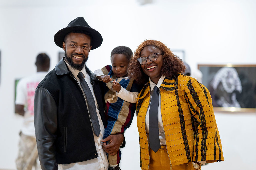 John Lekan in a black fedora smiling with a small child and a woman in a yellow striped outfit in the gallery