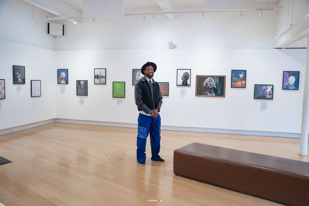 The white gallery at LCB Depot with John Lekan standing in a black fedora and blue patterned trousers among the framed works