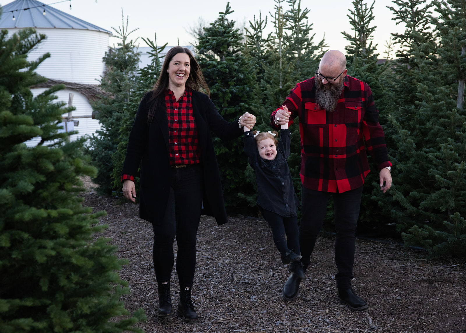 Parents holding their smiling daughter’s hands among Christmas trees during a family photo session