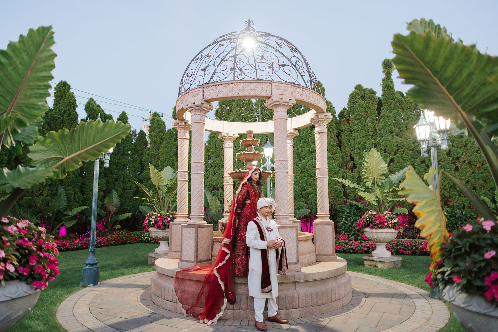 Bhavna and Ricky pose beside a stone gazebo and fountain, surrounded by garden foliage and pink flowers.