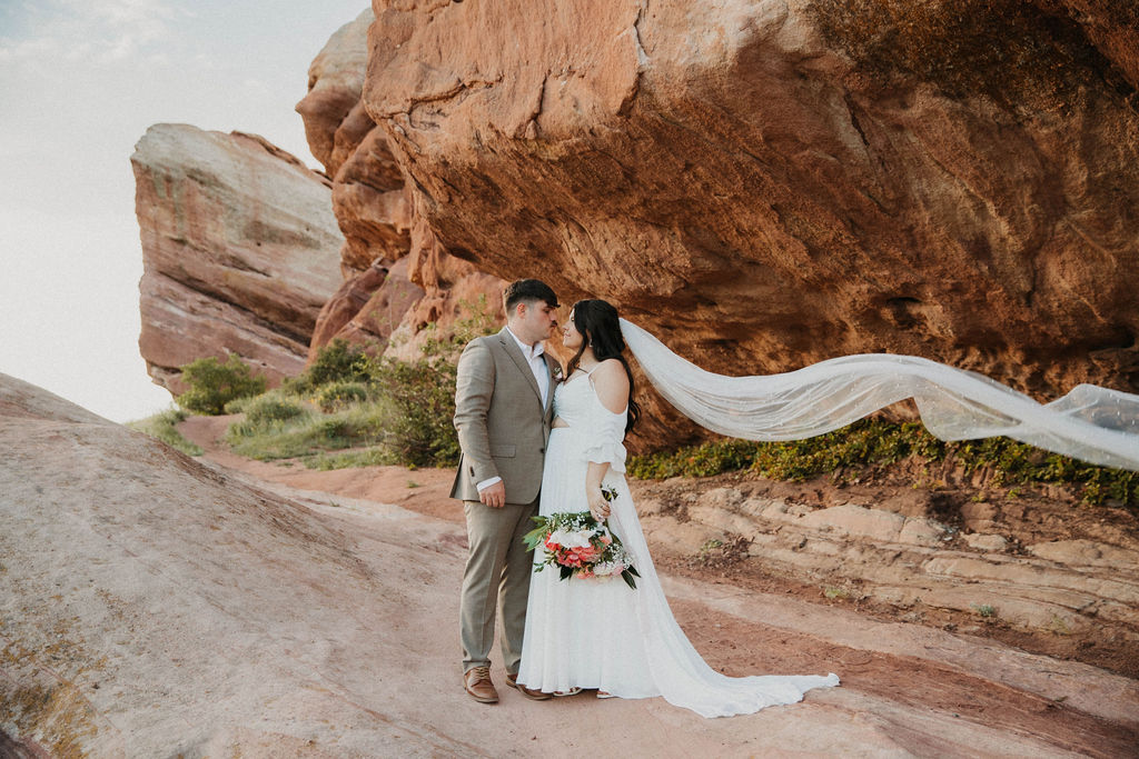 Bride and groom beneath red rock formations as her veil streams in the wind