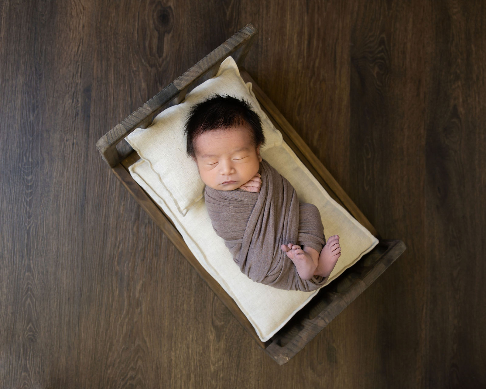 Sleeping newborn wrapped in taupe fabric and posed in a small wooden bed