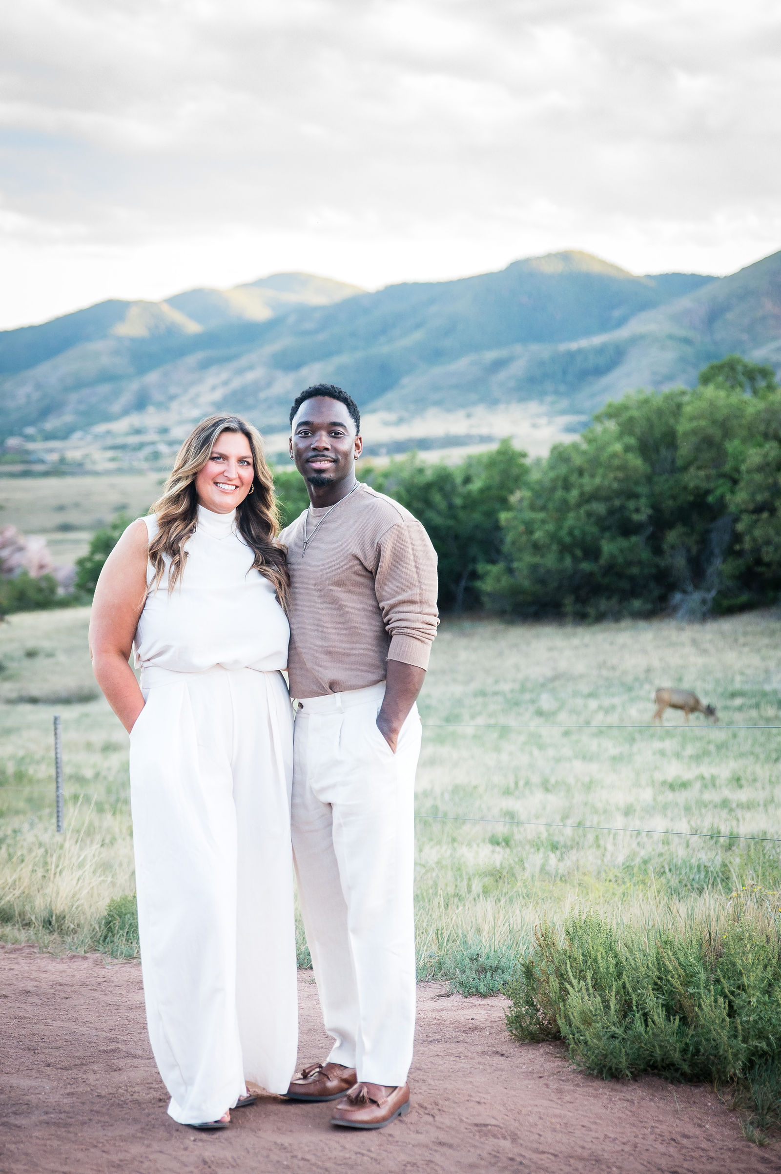 Engaged couple in a taupe sweater and white dress with the Colorado foothills behind them