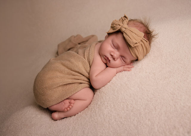 Sleeping newborn girl wrapped in warm neutral fabric with a bow during a posed studio newborn session