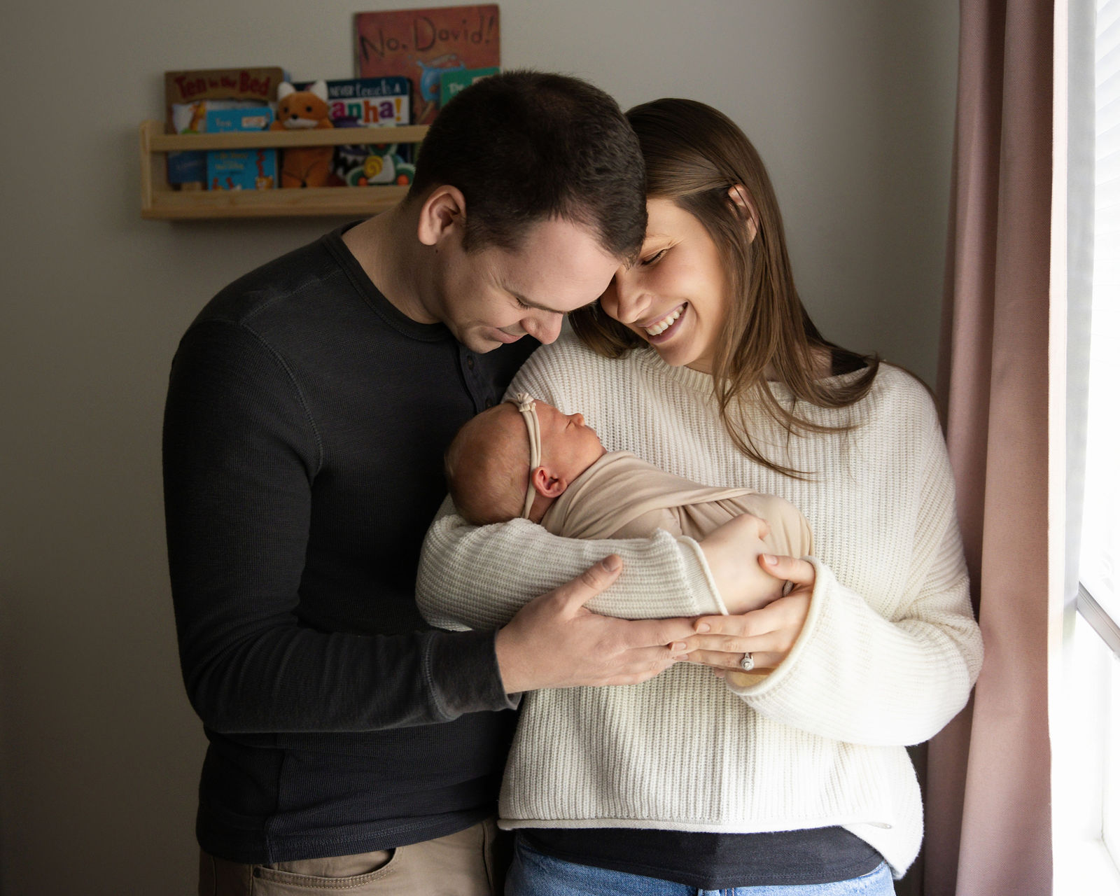 Parents standing together and smiling down at their newborn baby during an at-home newborn photography session