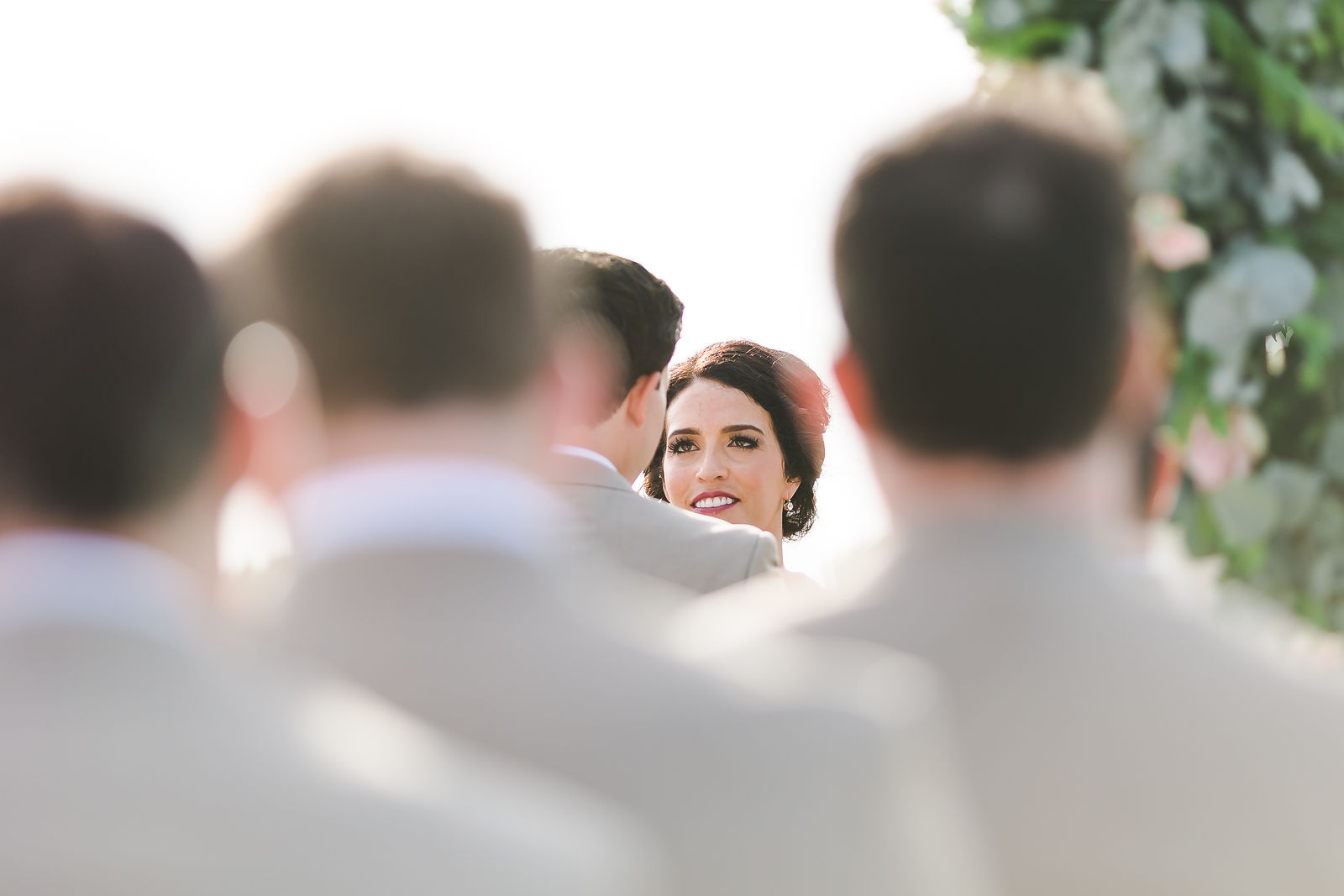 Bride smiling during the ceremony seen through guests, documented by the Kreate photo and film team