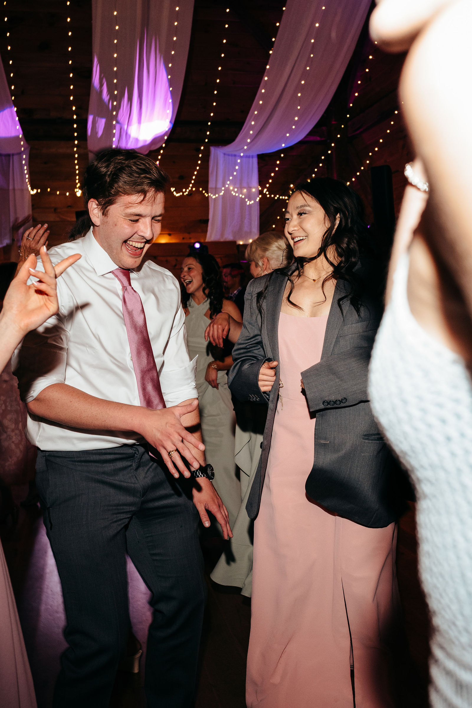 Guests laughing around a reception table with pink linens, grounds and gazebo visible through the barn windows at Dell-Lea