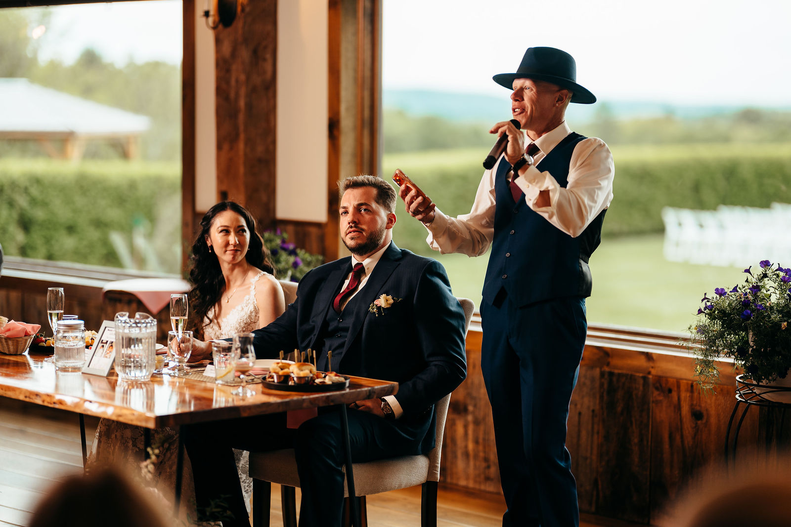 Guest giving a toast at the head table with Dell-Lea's green grounds visible through floor-to-ceiling windows