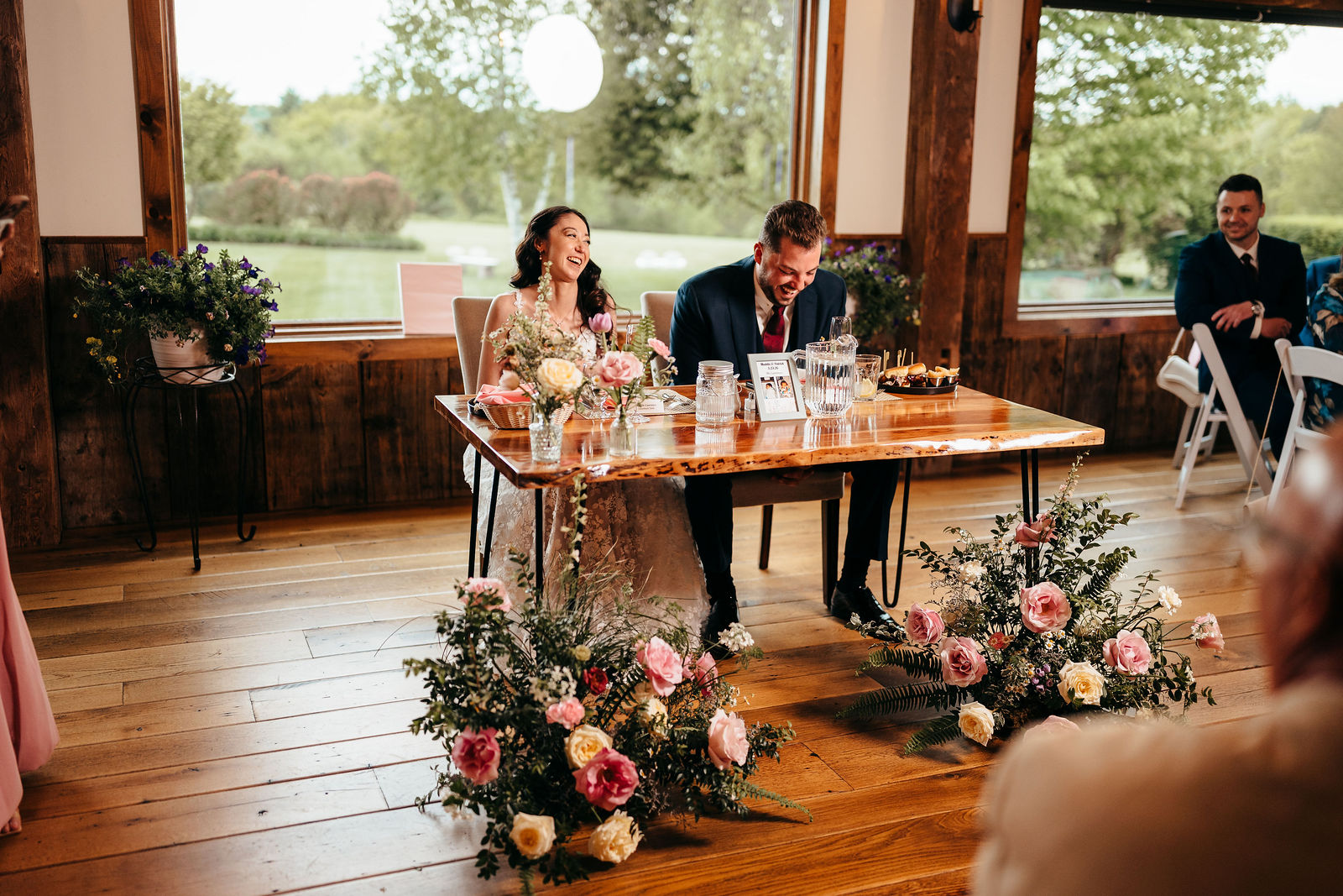 Couple laughing at their wooden sweetheart table with pink and white florals at Dell-Lea